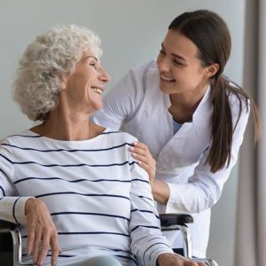 Healthcare professional smiling while assisting an elderly woman in a wheelchair, showcasing compassionate care and support in a social care setting.