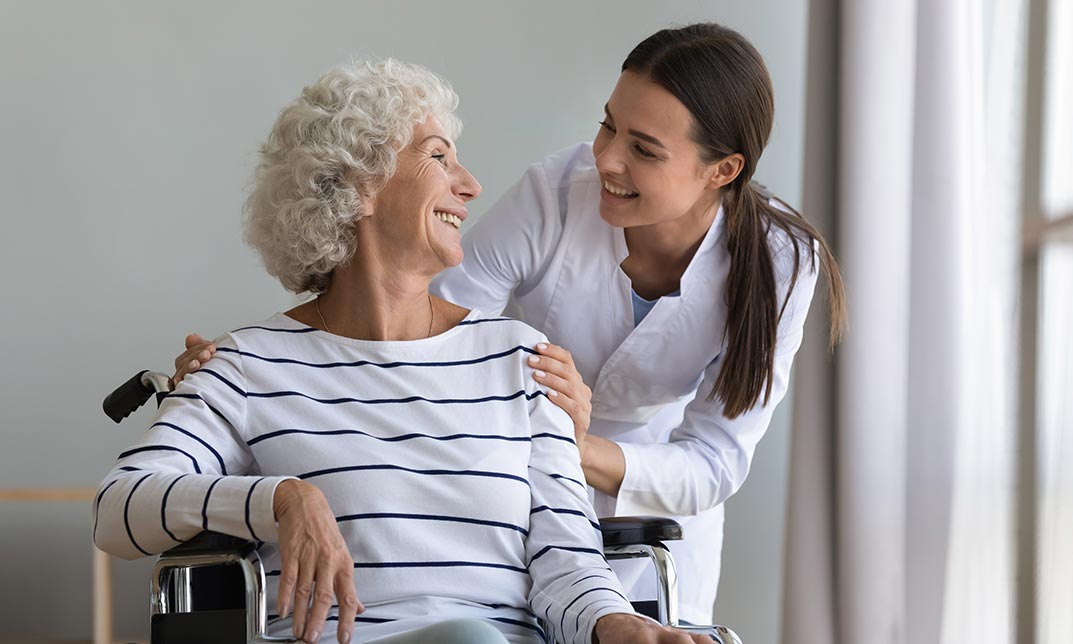 Healthcare professional smiling while assisting an elderly woman in a wheelchair, showcasing compassionate care and support in a social care setting.