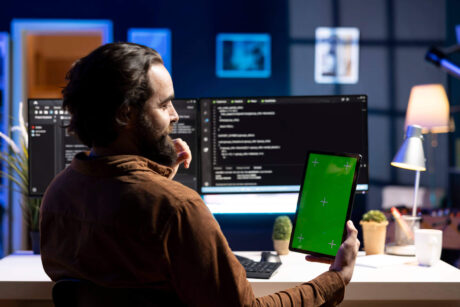 A developer sits at a desk with multiple screens displaying code while holding a tablet with a green screen, working on a Python GUI development project using Tkinter.