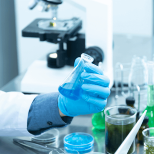 Close-up of a laboratory setup featuring a researcher holding a flask with a blue liquid, surrounded by petri dishes and glassware, with a microscope in the background.